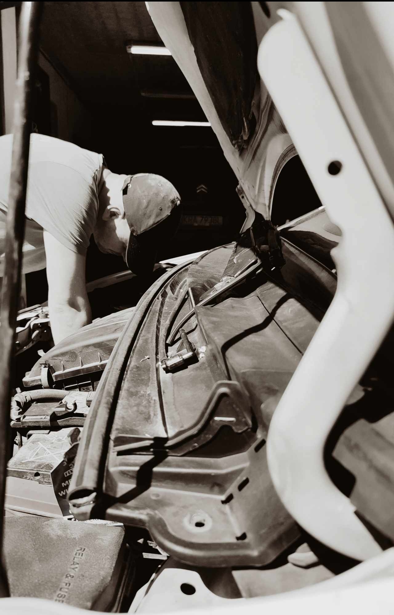 Czarno-białe zdjęcie samochodu z otwartą maską, gdzie mężczyzna w czapce pochyla się nad silnikiem, prawdopodobnie wykonując naprawę lub diagnostykę.