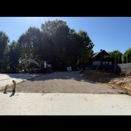 PROJAR - Przygotowany teren pod budowę z ubitym podłożem i widocznym w tle koparką. W tle drzewa i budynek.