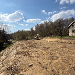 PROJAR - Obszerny teren po pracach ziemnych, z koparką kołową w oddali i domem po prawej, pod czystym, błękitnym niebem.