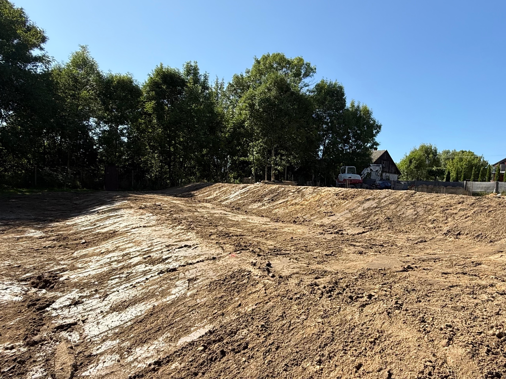 Niwelacja terenu: wyrównany teren z widocznymi śladami pracy maszyn, w tle drzewa i budynek. Błękitne niebo.