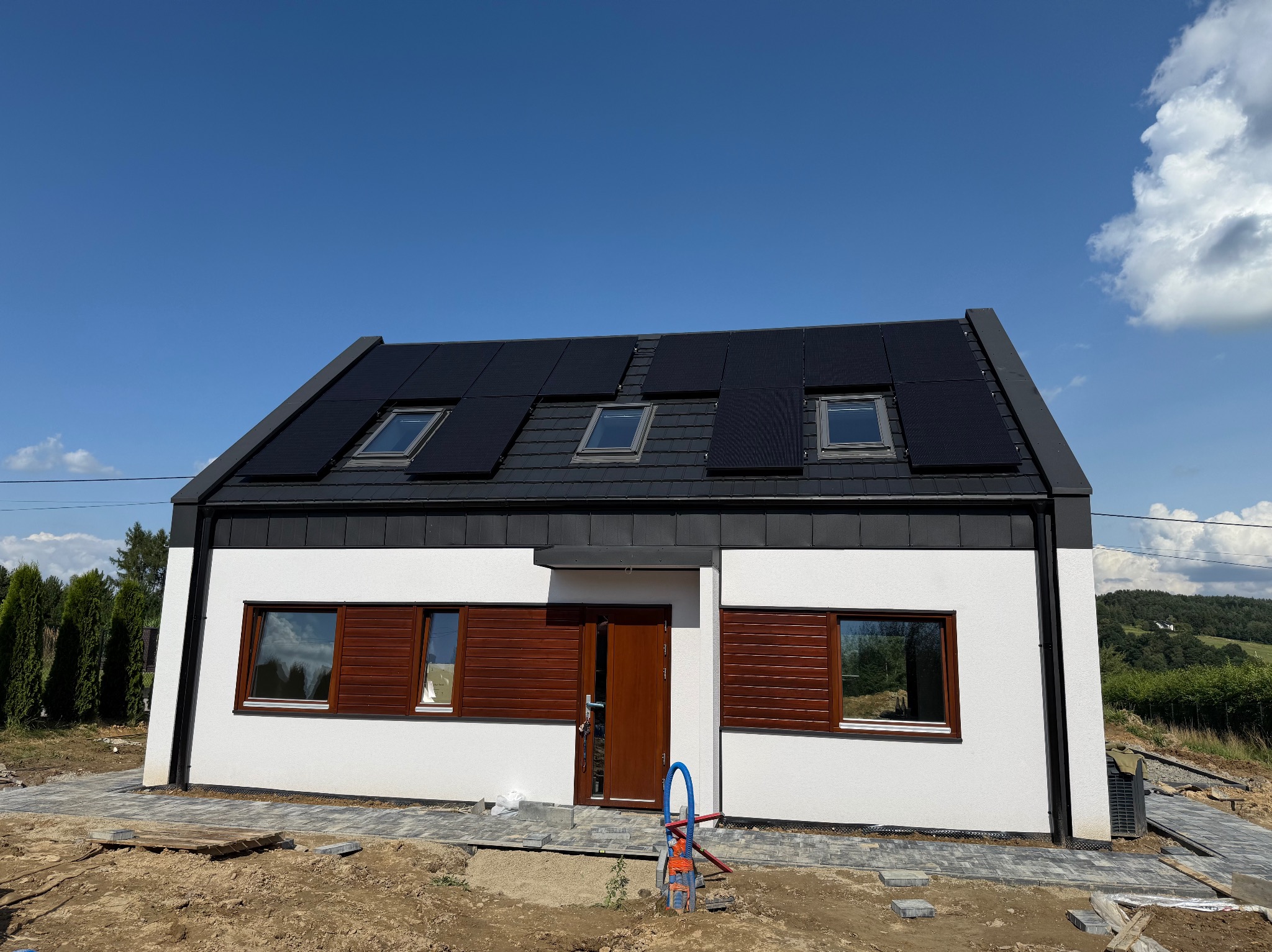 Biały dom z panelami słonecznymi na dachu i brązowymi oknami, widoczny fragment układanej kostki brukowej na podwórku, błękitne niebo w tle.