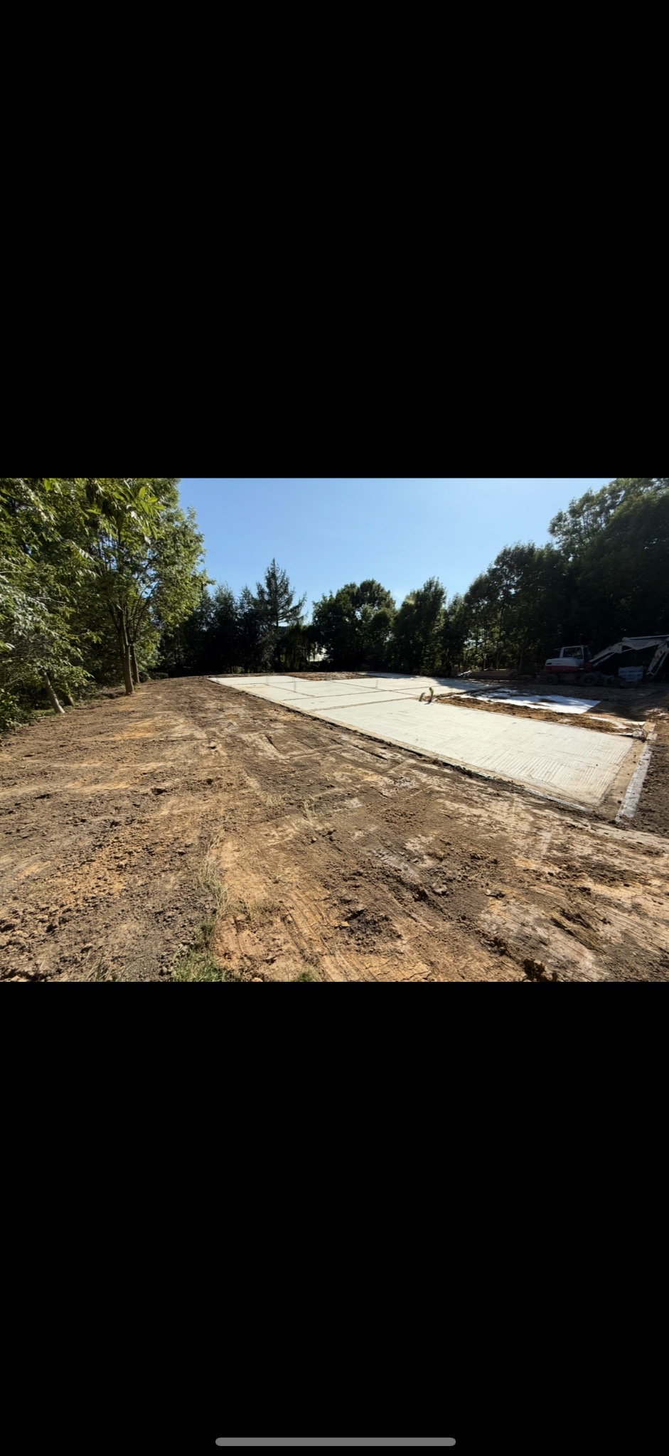 Przygotowany teren pod budowę z wylanym fundamentem i widoczną koparką w tle, otoczony drzewami w słoneczny dzień. Widok z poziomu gruntu.