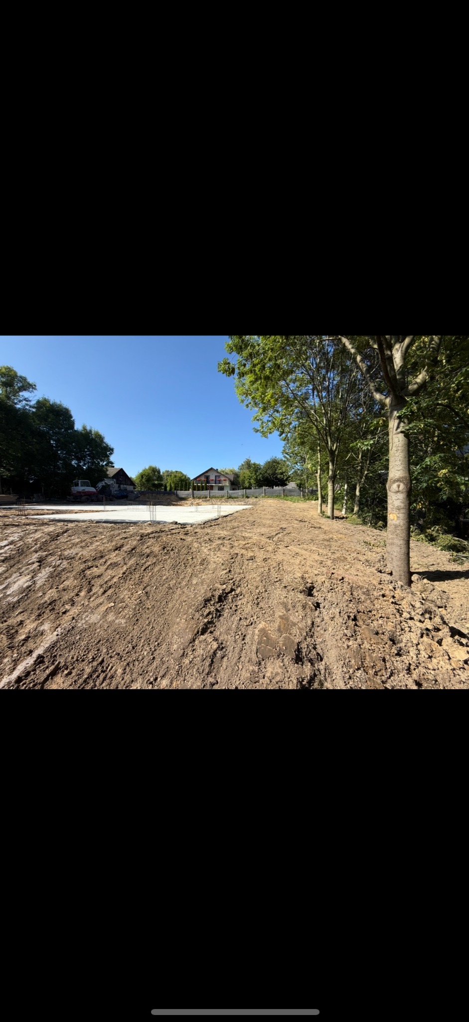 Przygotowany teren pod budowę domu z widocznym zbrojeniem i fundamentami. W tle domy jednorodzinne i zieleń drzew.
