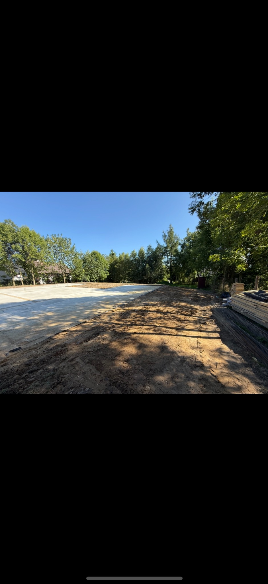 Przygotowany teren pod budowę z częściowo wylanym betonem, widoczne drzewa w tle i składowane materiały budowlane.