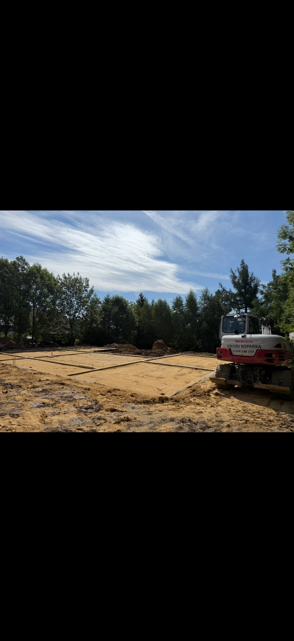 Przygotowany wykop pod fundamenty z widoczną konstrukcją szalunkową i mini koparką Takeuchi z boku, na tle drzew i nieba. Widoczne ślady pracy ziemnej.