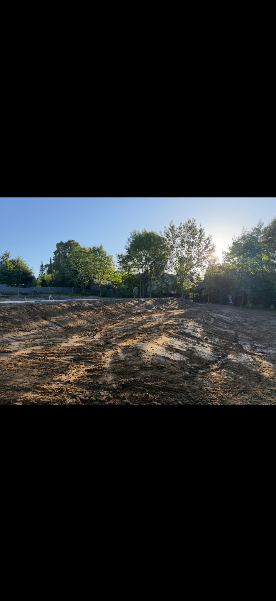 Ukształtowany teren pod budowę w słoneczny dzień. Widoczne świeże ślady maszyn, przygotowana ziemia pod dalsze prace budowlane, drzewa w tle.