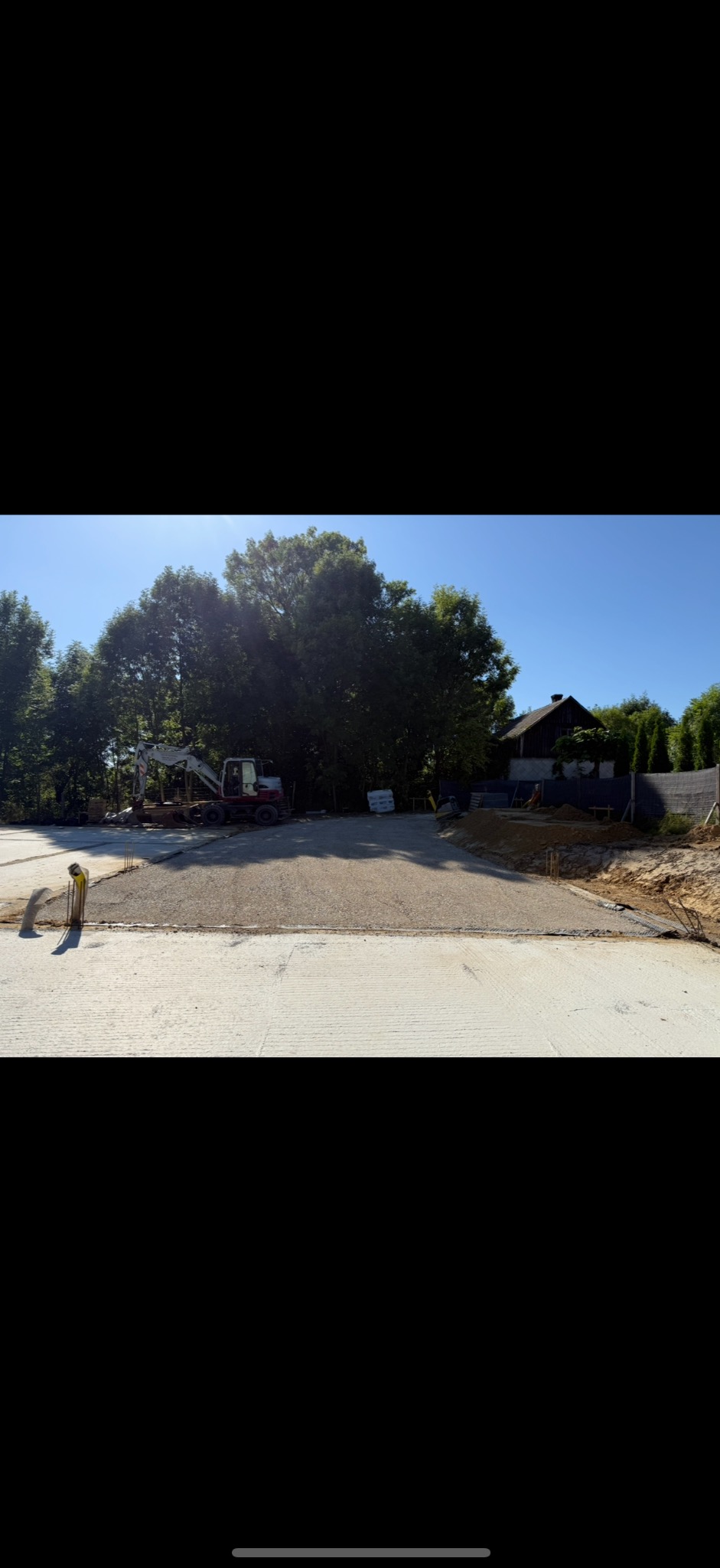 Przygotowany teren pod budowę z ubitym podłożem i widocznym w tle koparką. W tle drzewa i budynek.