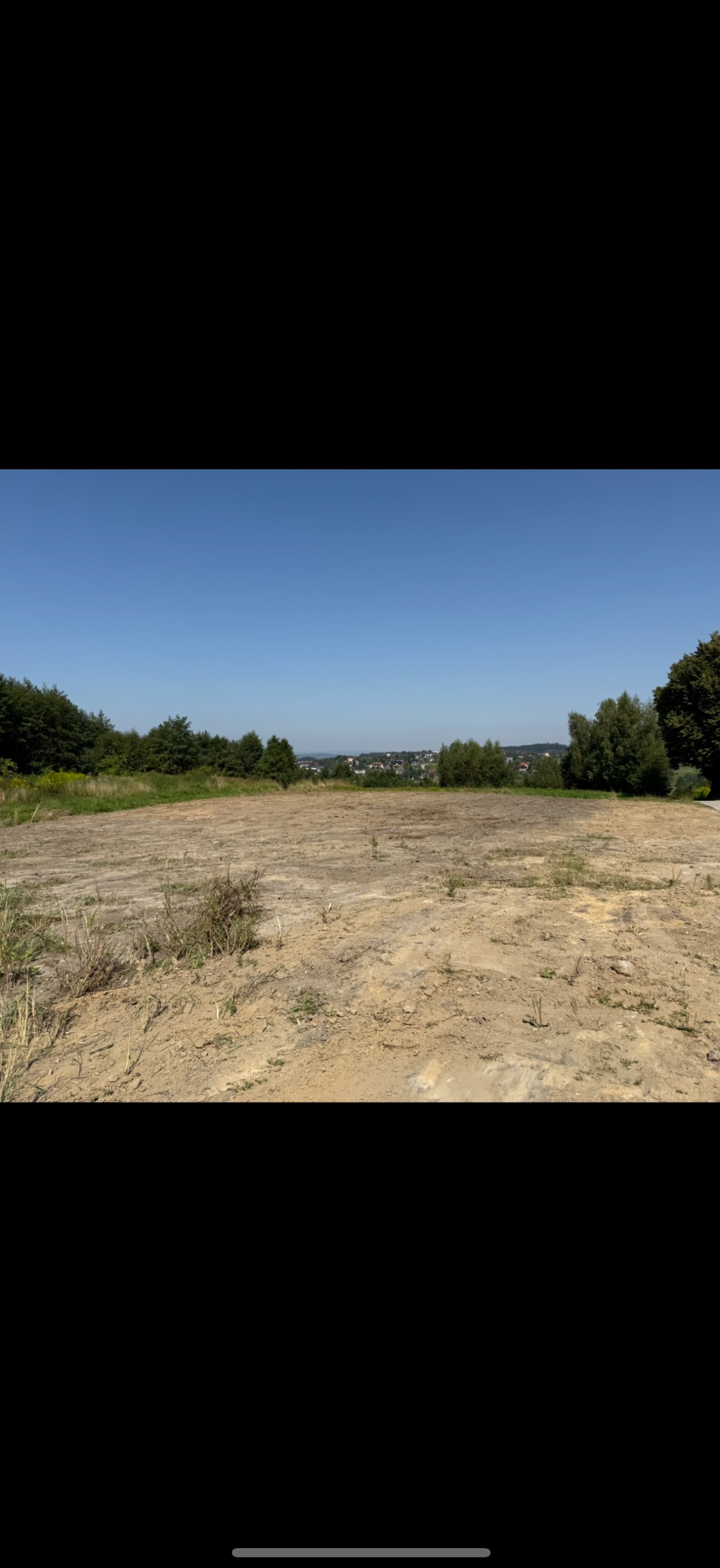 Wyrównany teren pod budowę z widokiem na zabudowania w oddali. Na pierwszym planie piasek, w tle zieleń drzew i błękitne niebo.