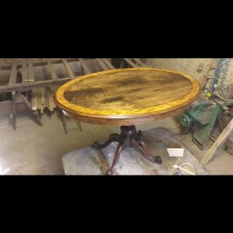 Sebastian Banaszek - Owalny, drewniany stolik po renowacji, z widocznym nowym fornirem w odcieniach orzecha i dębu, stojący w warsztacie na tle maszyn stolarskich.