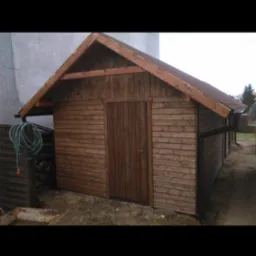 Drewniana szopa ogrodowa z brązowymi deskami, widoczne drzwi i rynna, obok leży zwinięty wąż ogrodowy.