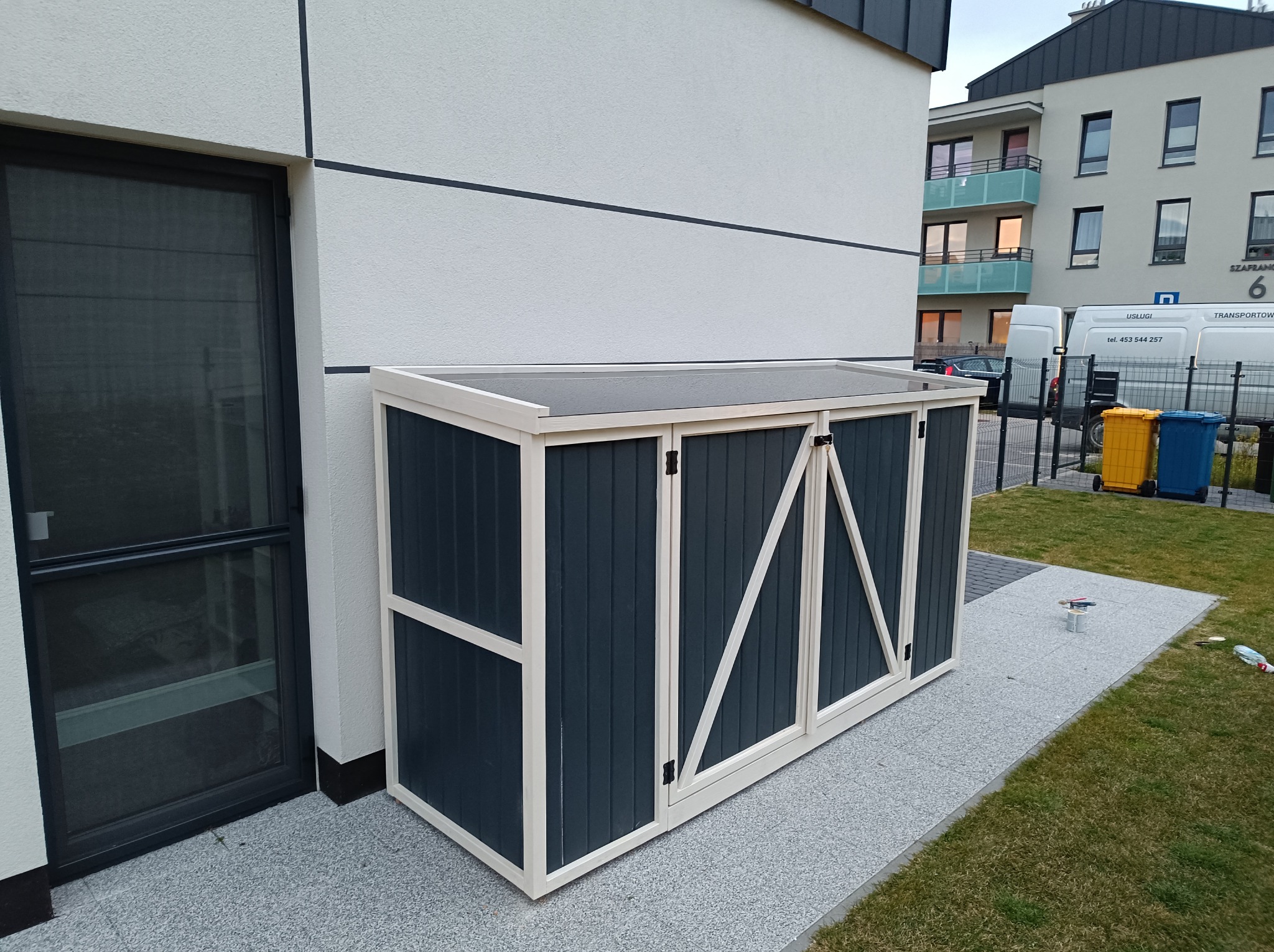 Zewnętrzna szafa ogrodowa w kolorze grafitowym z jasnymi obramowaniami, ustawiona na szarym chodniku z kostki, przy ścianie budynku o jasnej elewacji. Na chodniku widoczne ślady farby i pędzle.