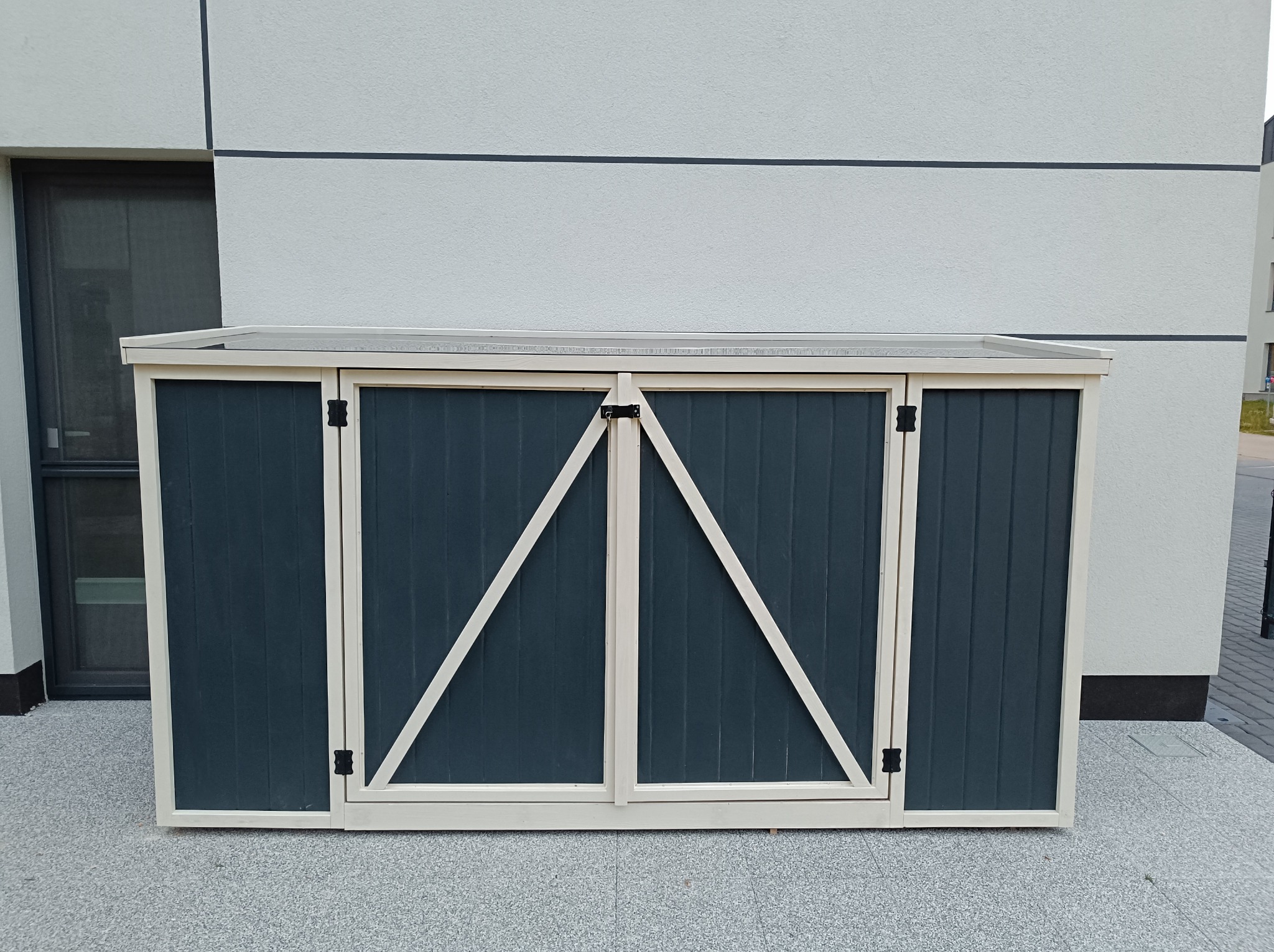 Trzydrzwiowa, ciemnoszara skrzynia ogrodowa z beżowymi ramami i ukośnymi wzmocnieniami, stojąca na szarym, kamiennym podłożu przed jasnym budynkiem.