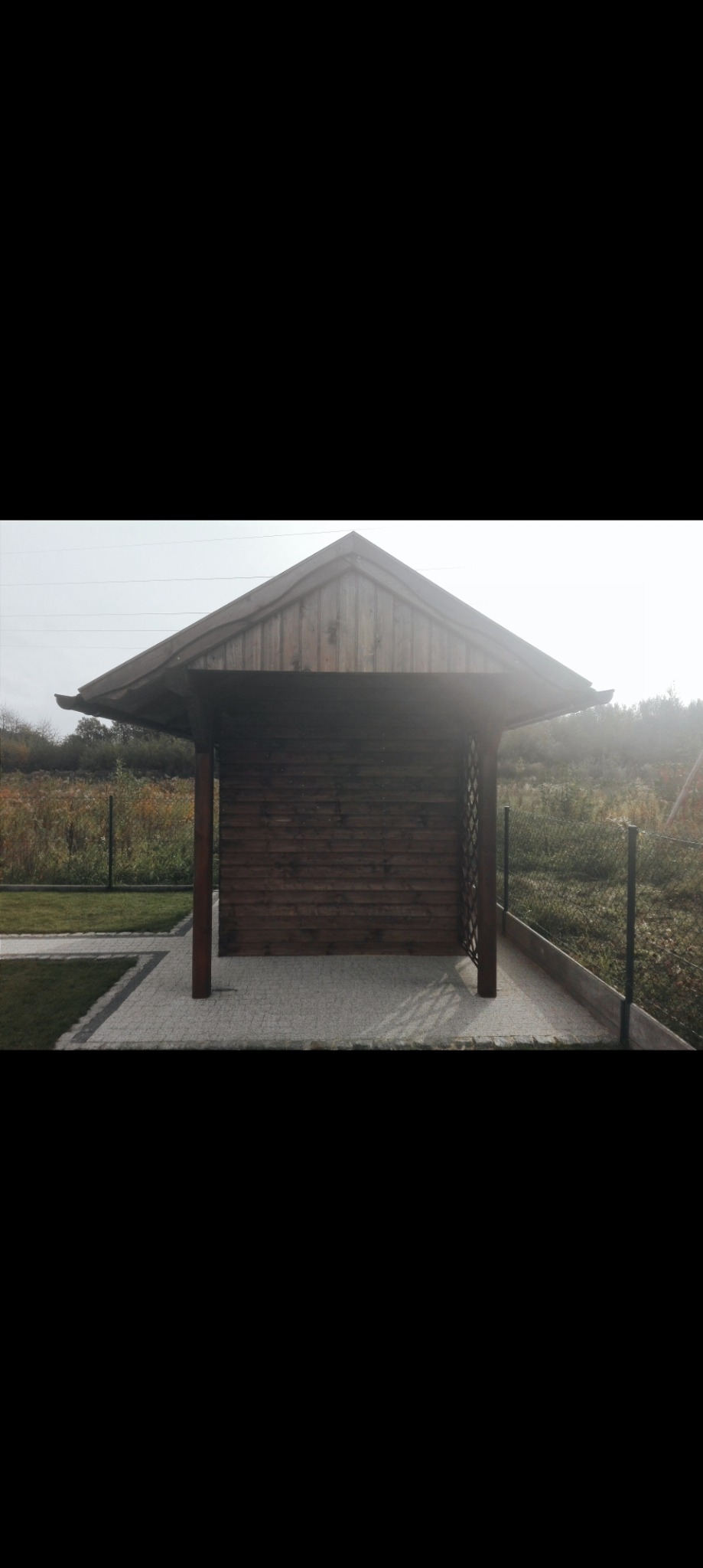 Drewniana altana z kratownicą w Pasłęku, otoczona zielenią, z kostką brukową i niskim ogrodzeniem, sfotografowana w pochmurny dzień.