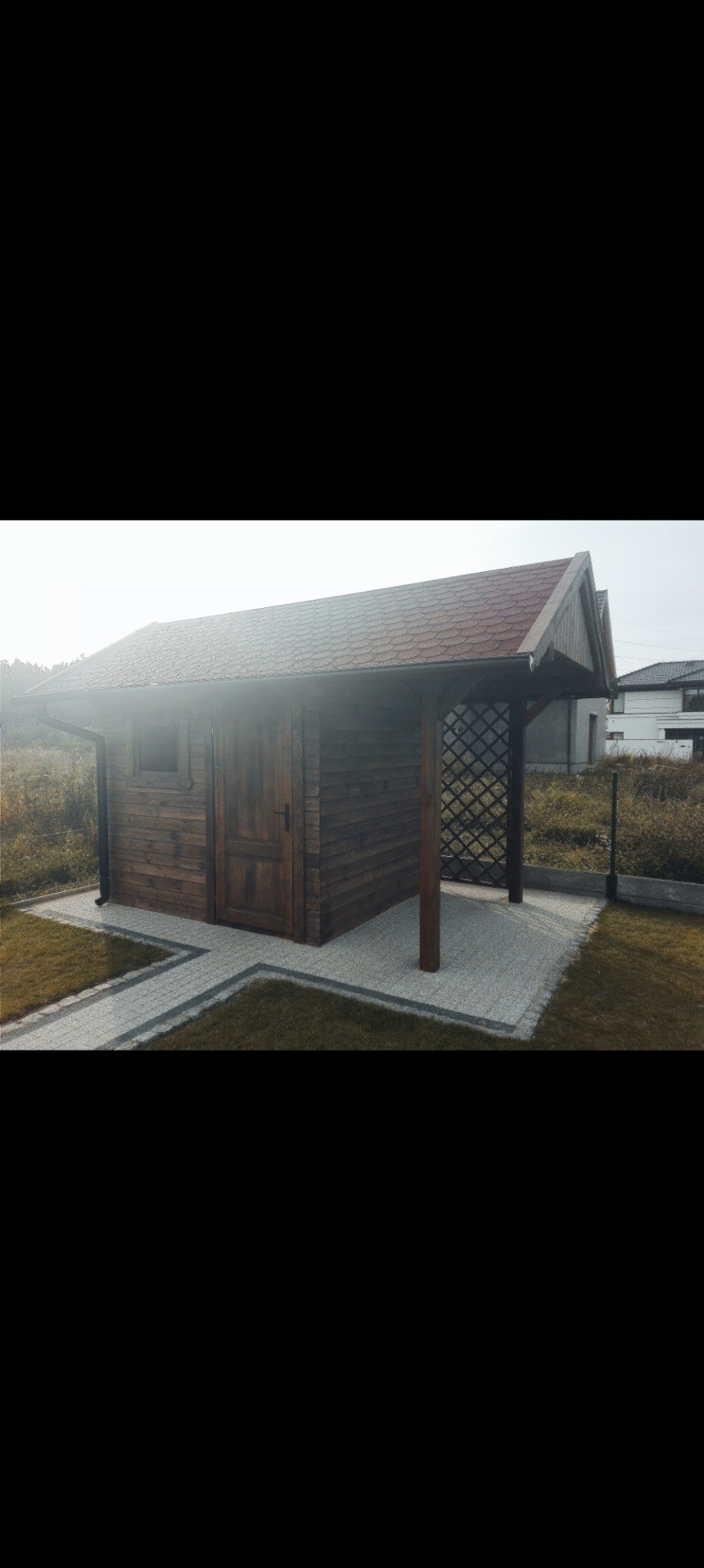 Drewniana altana z kratownicą, pokryta czerwoną dachówką, z widoczną rynną, otoczona trawnikiem i ścieżką z kostki brukowej, na tle zabudowań.