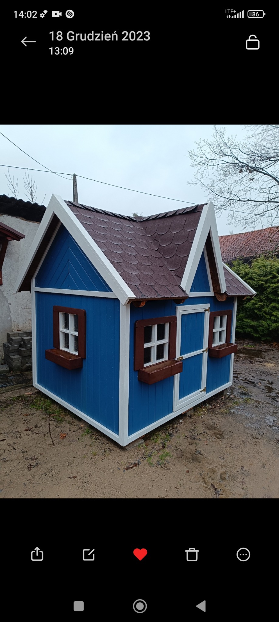 Niebieski domek zabaw dla dzieci z brązowym dachem, białymi ramami i okiennicami oraz doniczkami pod oknami, stojący na mokrej ziemi.