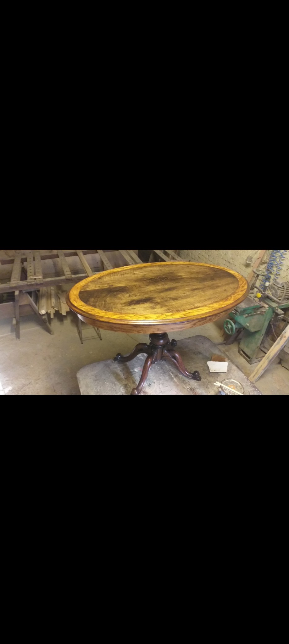 Owalny, drewniany stolik po renowacji, z widocznym nowym fornirem w odcieniach orzecha i dębu, stojący w warsztacie na tle maszyn stolarskich.