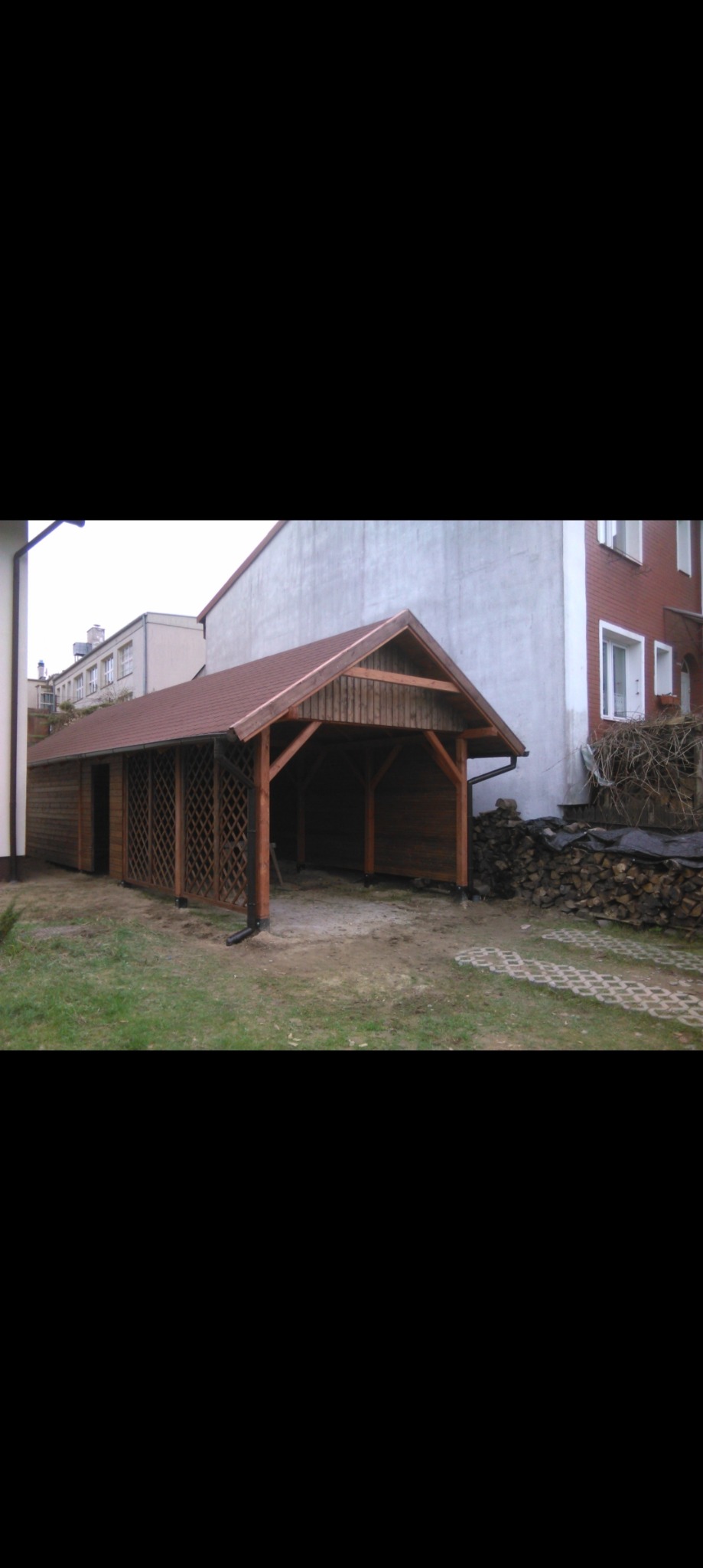 Długa, drewniana altana z kratownicowymi ścianami bocznymi i brązowym dachem, przylegająca do budynku mieszkalnego, z ułożonym drewnem opałowym w tle.