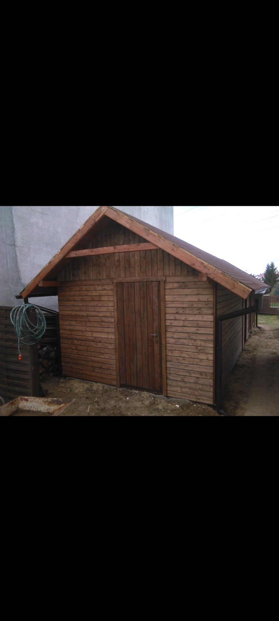 Drewniana szopa ogrodowa z brązowymi deskami, widoczne drzwi i rynna, obok leży zwinięty wąż ogrodowy.