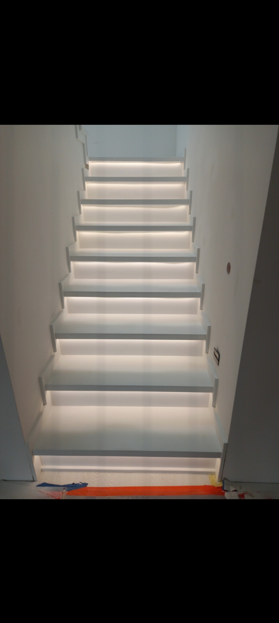 Białe schody z podświetleniem LED na każdym stopniu, widoczne zabezpieczenia podłogi na dole schodów.