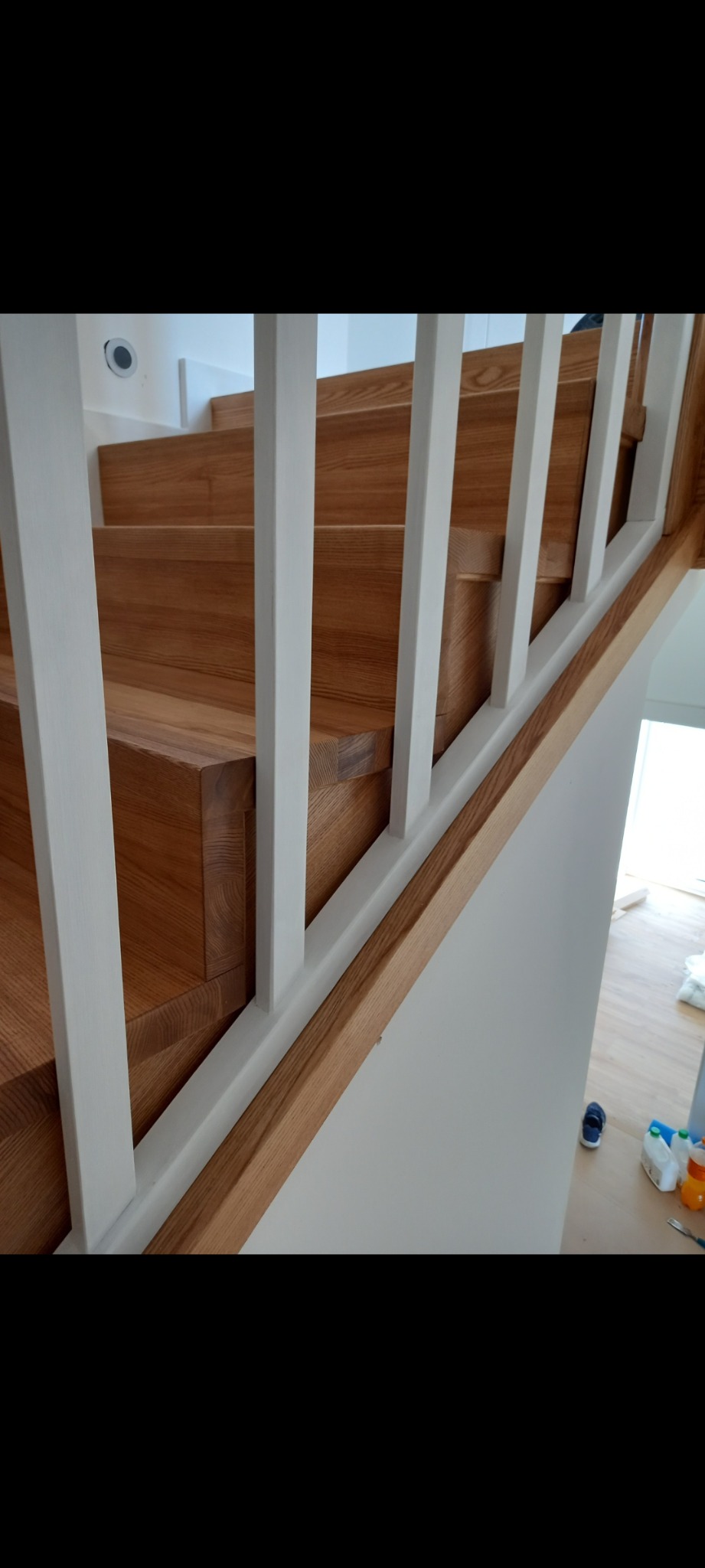 Nowoczesne schody drewniane z białymi balustradami, widok z dołu, na podłodze widoczne akcesoria malarskie i dziecięce buty.