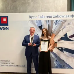 Dwie osoby, mężczyzna w garniturze trzymający statuetkę i kobieta w czarnej sukience trzymająca certyfikat na tle baneru WGN z hasłem 'Bycie Liderem zobowiązuje'.