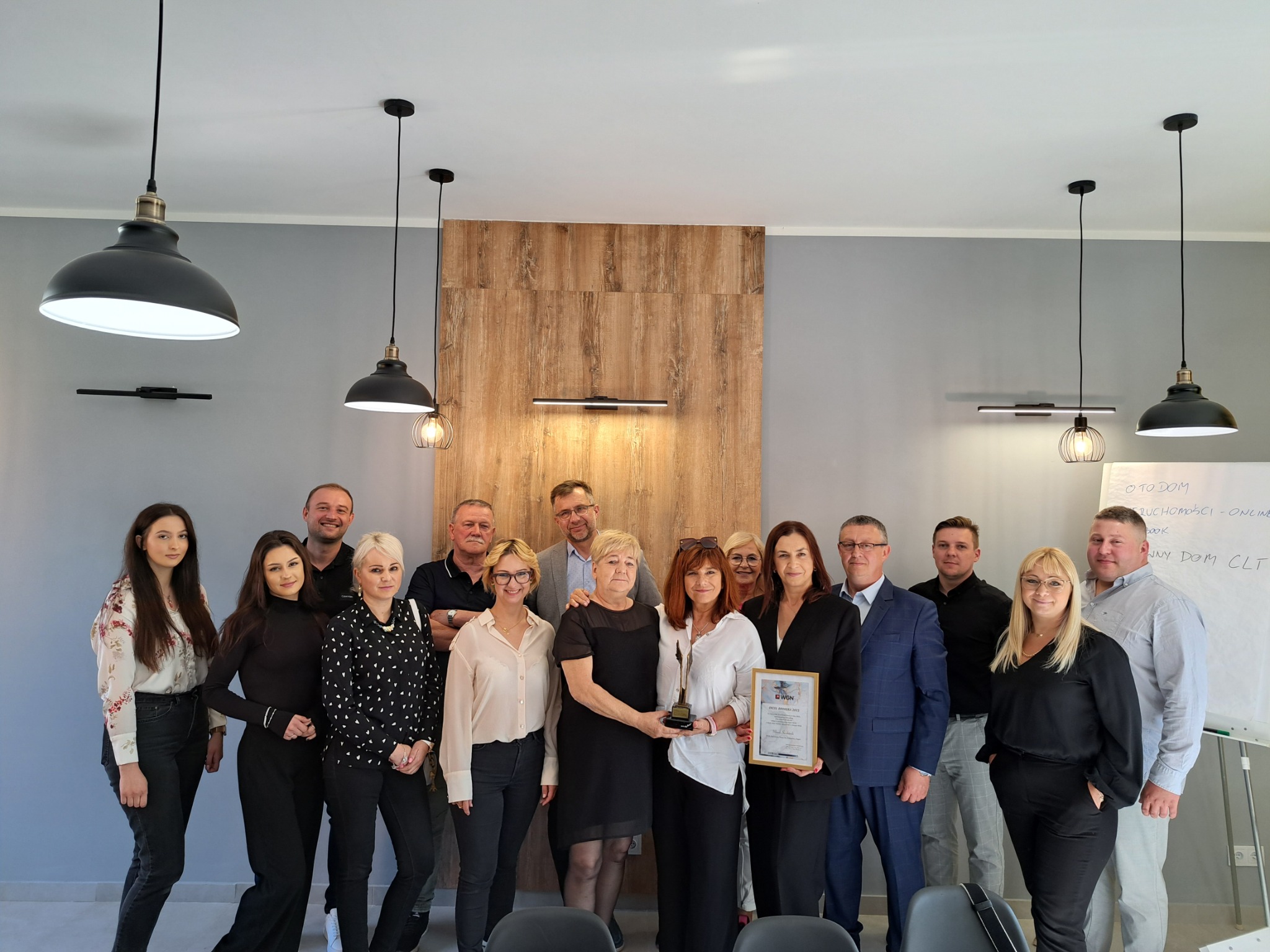 Zespół pracowników biura nieruchomości pozuje z nagrodą i certyfikatem w nowoczesnym wnętrzu z drewnianą ścianą akcentującą i designerskimi lampami wiszącymi.