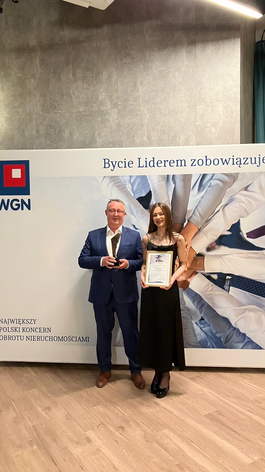 Dwie osoby, mężczyzna w garniturze trzymający statuetkę i kobieta w czarnej sukience trzymająca certyfikat na tle baneru WGN z hasłem 'Bycie Liderem zobowiązuje'.