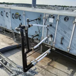 Instalacja rur na dachu budynku z widocznym systemem wentylacyjnym i elementami mocującymi. Rury czarne i białe, złącza metalowe, tło stanowi błękitne niebo.