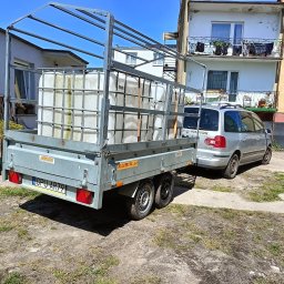 Pogotowie hydrauliczne wod-kan - Przyczepa z pojemnikami na wodę, podłączona do samochodu osobowego. Widoczne budynki w tle, dzień słoneczny.