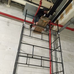 Pogotowie hydrauliczne wod-kan - Pracownik na rusztowaniu łączy rury kanalizacyjne wewnątrz budynku. Widoczne czarne rury, czerwona instalacja przeciwpożarowa i surowy mur z białej cegły.