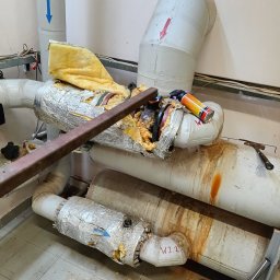 Pogotowie hydrauliczne wod-kan - Uszkodzona instalacja rur z izolacją, narzędzia i ślady wycieków. Rury oznaczone strzałkami i napisem 'W2.2'. Widoczne elementy demontażu.