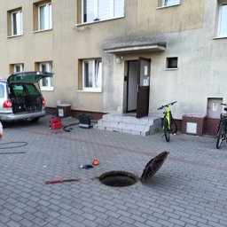 Pogotowie hydrauliczne wod-kan - Otwarty właz kanalizacyjny i sprzęt hydrauliczny na chodniku we Władysławowie. W tle budynek mieszkalny i samochód z otwartym bagażnikiem.
