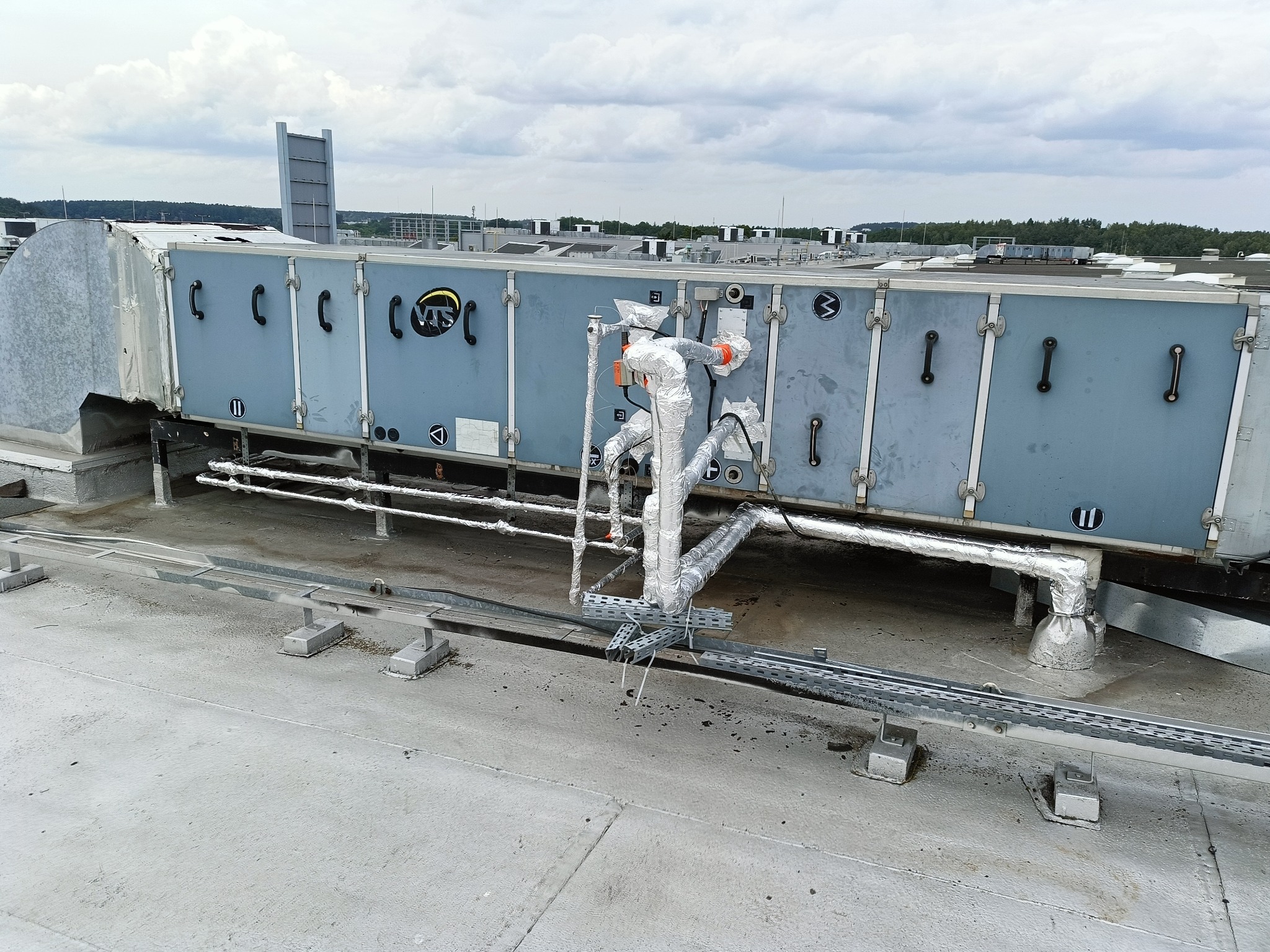 Instalacja wentylacyjna na dachu budynku, z widocznymi rurami owiniętymi folią aluminiową i elementami konstrukcyjnymi. Widok z góry, pochmurne niebo w tle.