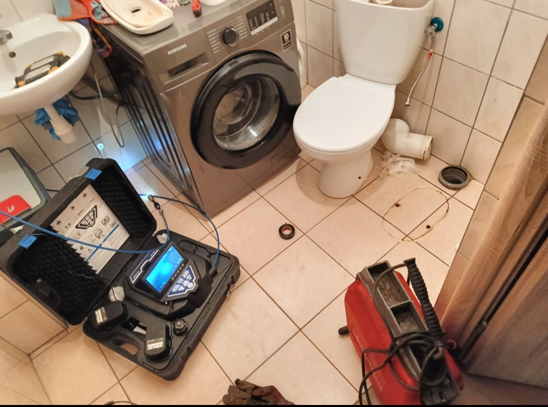 Sprzęt do inspekcji kanalizacji w łazience z pralką Samsung i toaletą. Widoczne narzędzia hydrauliczne i otwór w podłodze sugerują prace związane z udrażnianiem.