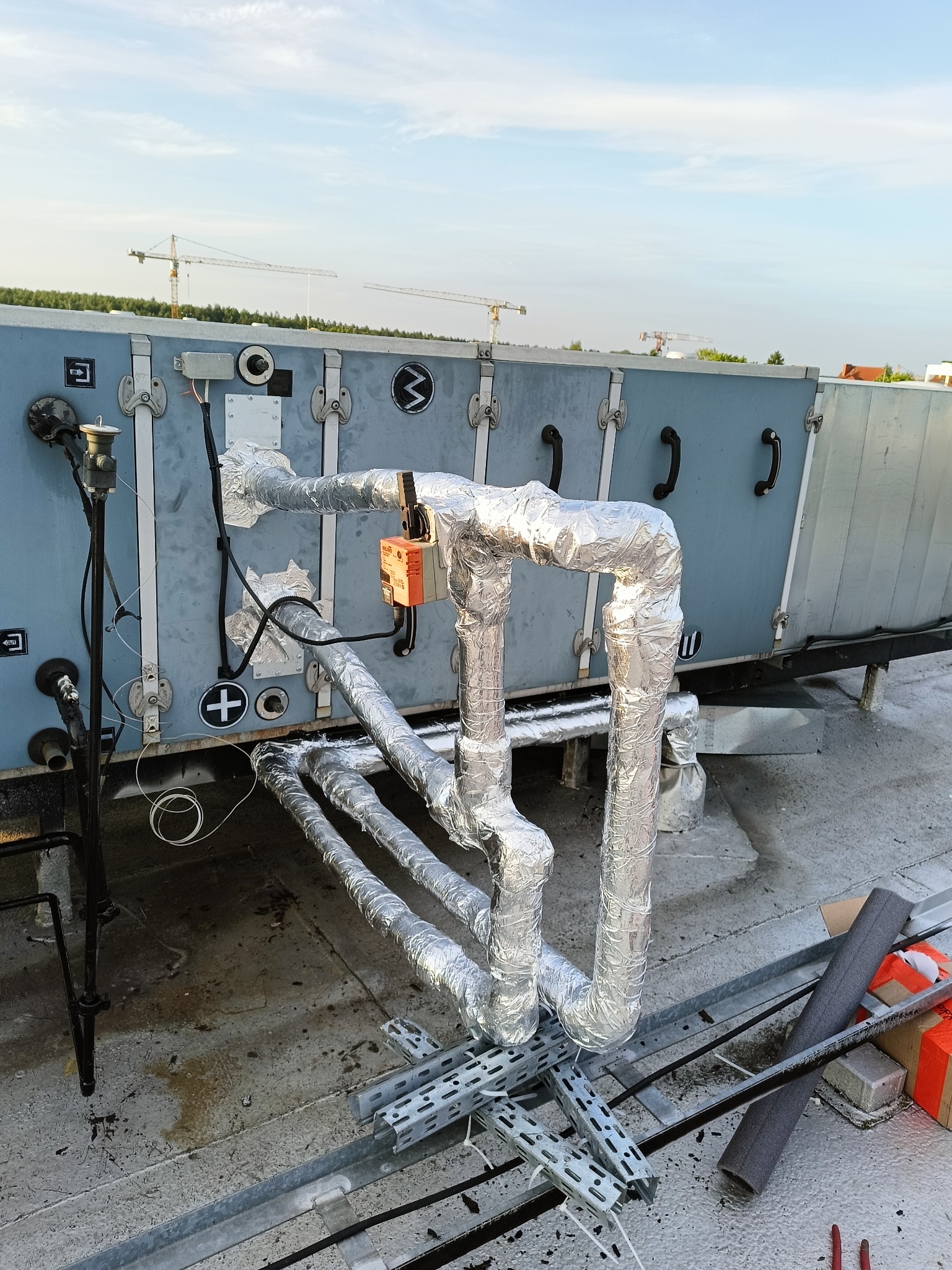 Instalacja przemysłowa na dachu budynku z izolowanymi rurami, skrzynką sterowniczą i elementami montażowymi na tle nieba z żurawiami budowlanymi.