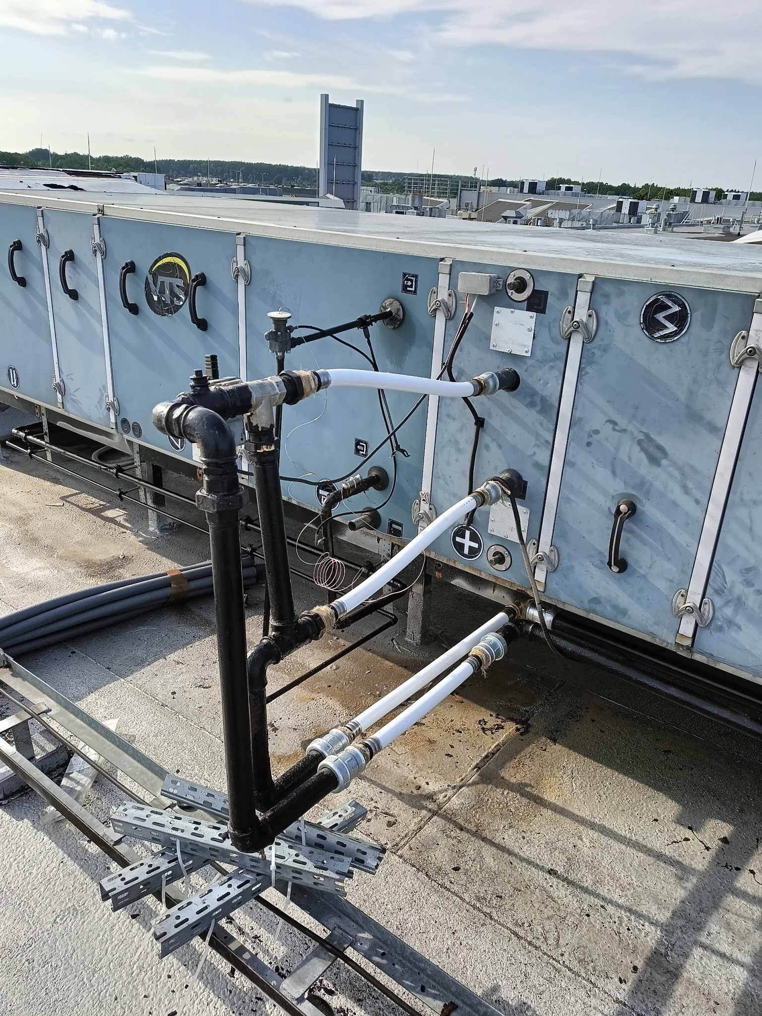 Instalacja rur na dachu budynku z widocznym systemem wentylacyjnym i elementami mocującymi. Rury czarne i białe, złącza metalowe, tło stanowi błękitne niebo.