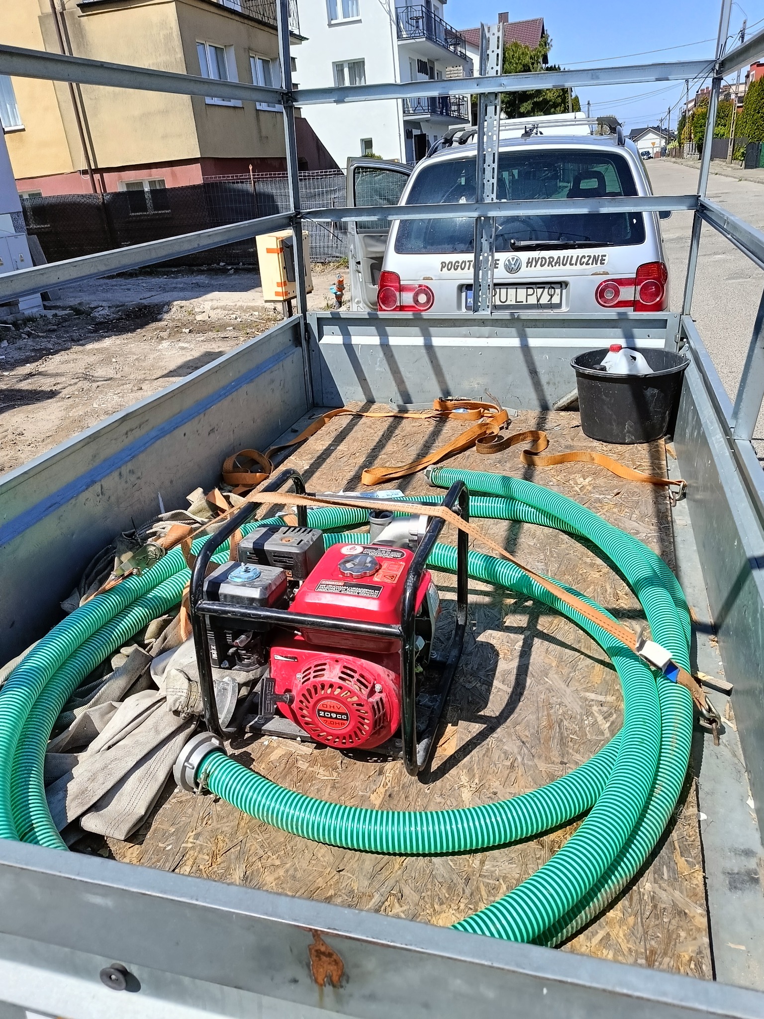 Przygotowany sprzęt hydrauliczny w przyczepie: czerwona pompa spalinowa, zwinięte węże ssące, wiadro i pasy transportowe. W tle samochód z napisem 'Pogotowie Hydrauliczne'.