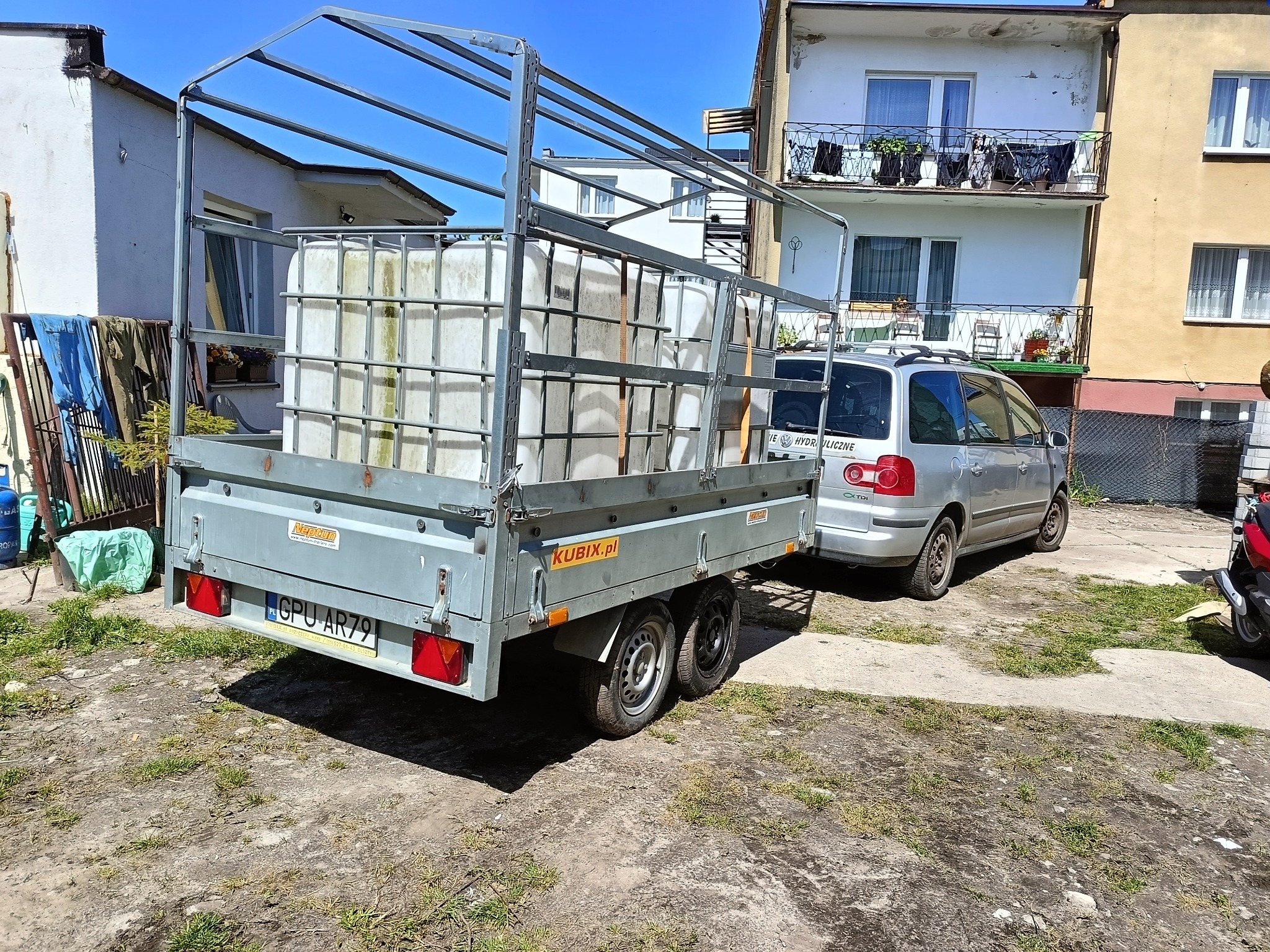 Przyczepa z pojemnikami na wodę, podłączona do samochodu osobowego. Widoczne budynki w tle, dzień słoneczny.