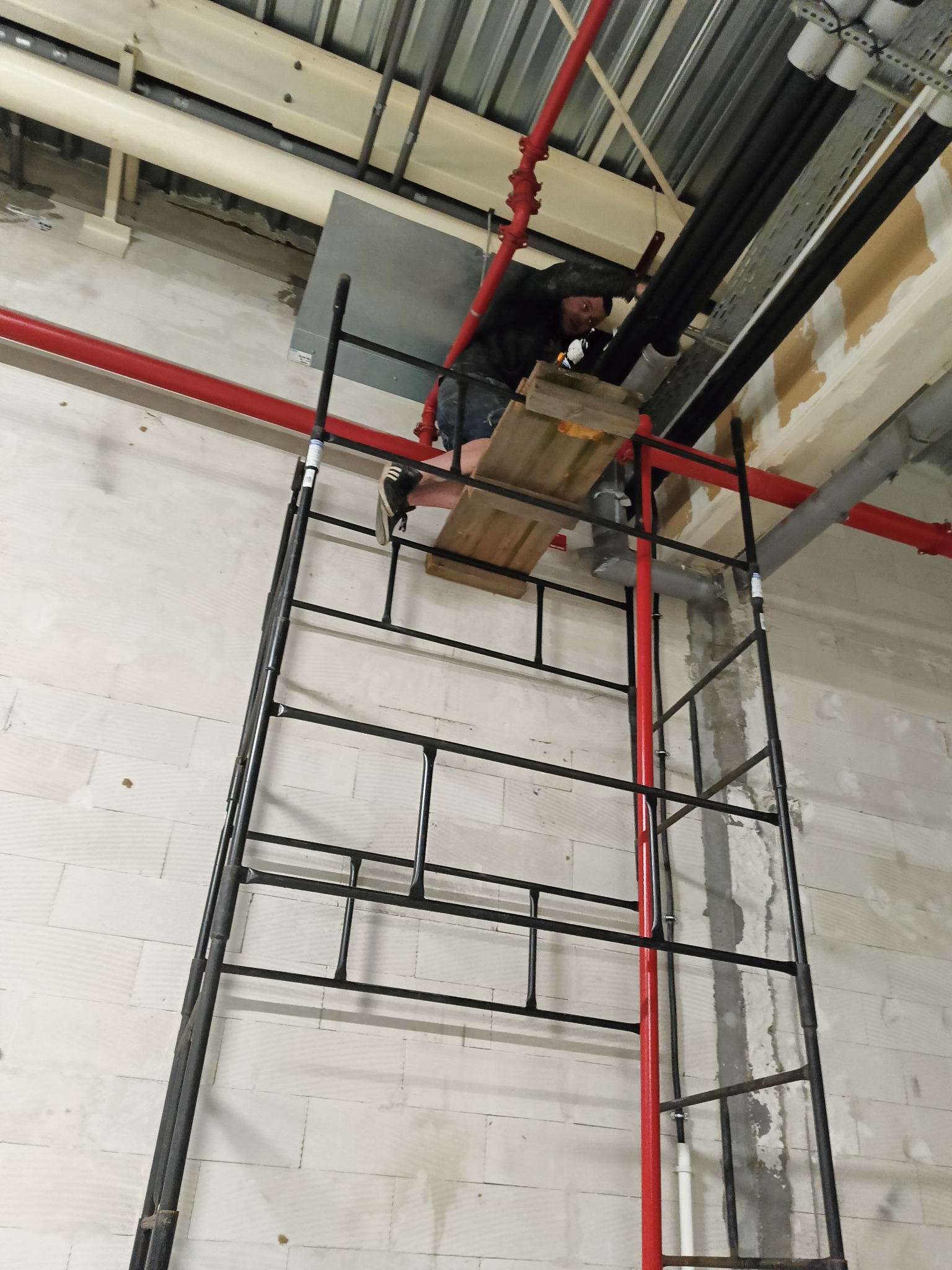 Pracownik na rusztowaniu łączy rury kanalizacyjne wewnątrz budynku. Widoczne czarne rury, czerwona instalacja przeciwpożarowa i surowy mur z białej cegły.
