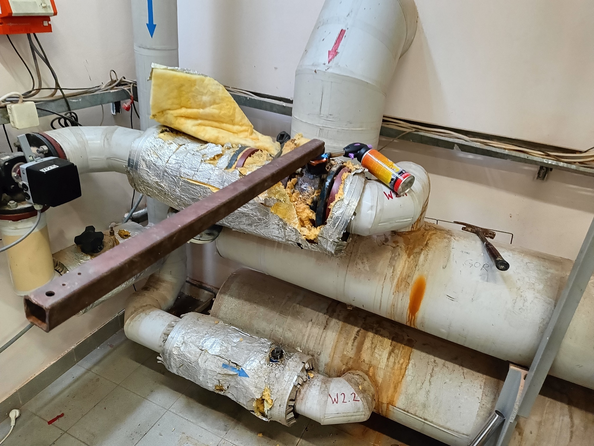 Uszkodzona instalacja rur z izolacją, narzędzia i ślady wycieków. Rury oznaczone strzałkami i napisem 'W2.2'. Widoczne elementy demontażu.