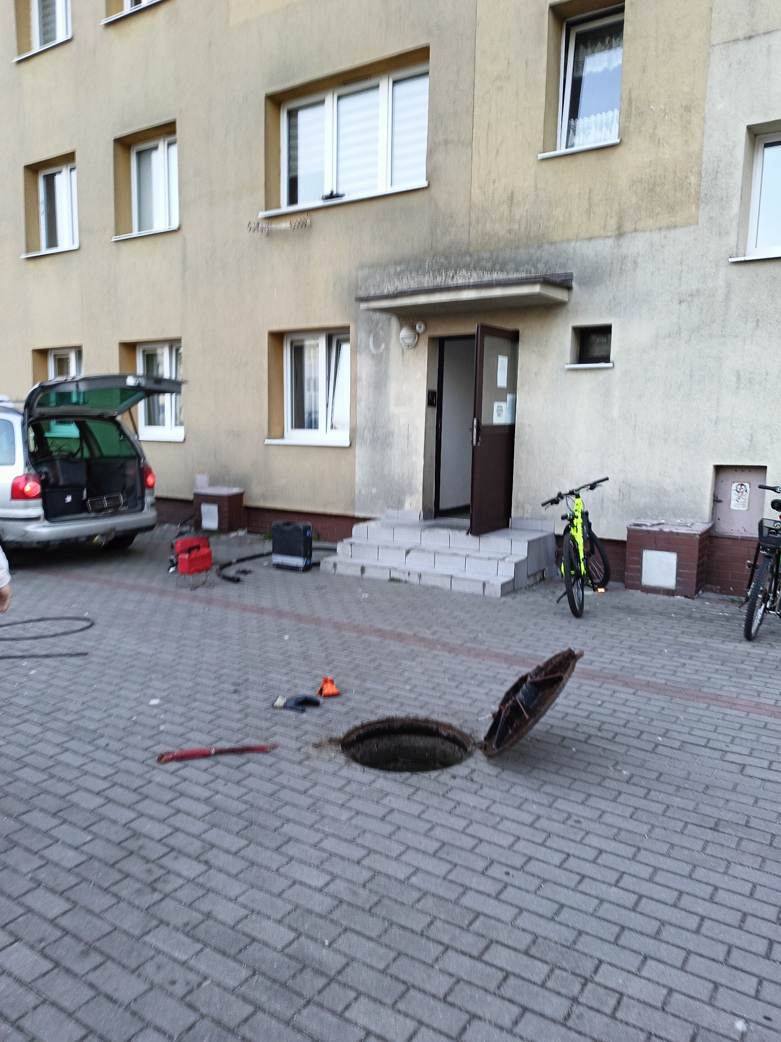 Otwarty właz kanalizacyjny i sprzęt hydrauliczny na chodniku we Władysławowie. W tle budynek mieszkalny i samochód z otwartym bagażnikiem.