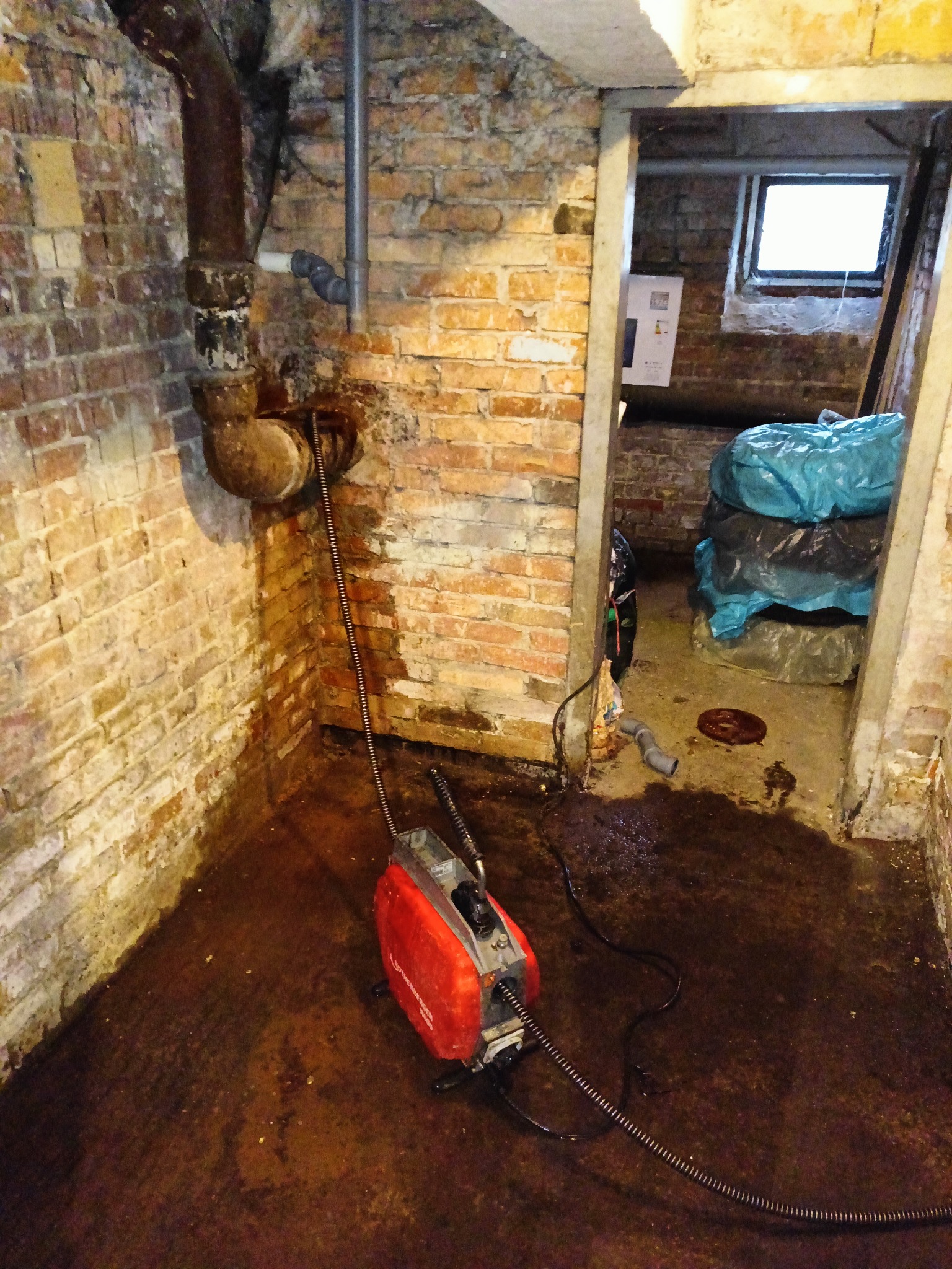 Czerwone urządzenie do udrażniania rur z wężem w brudnej piwnicy z ceglanymi ścianami. W tle otwarte drzwi do drugiego pomieszczenia z workami.