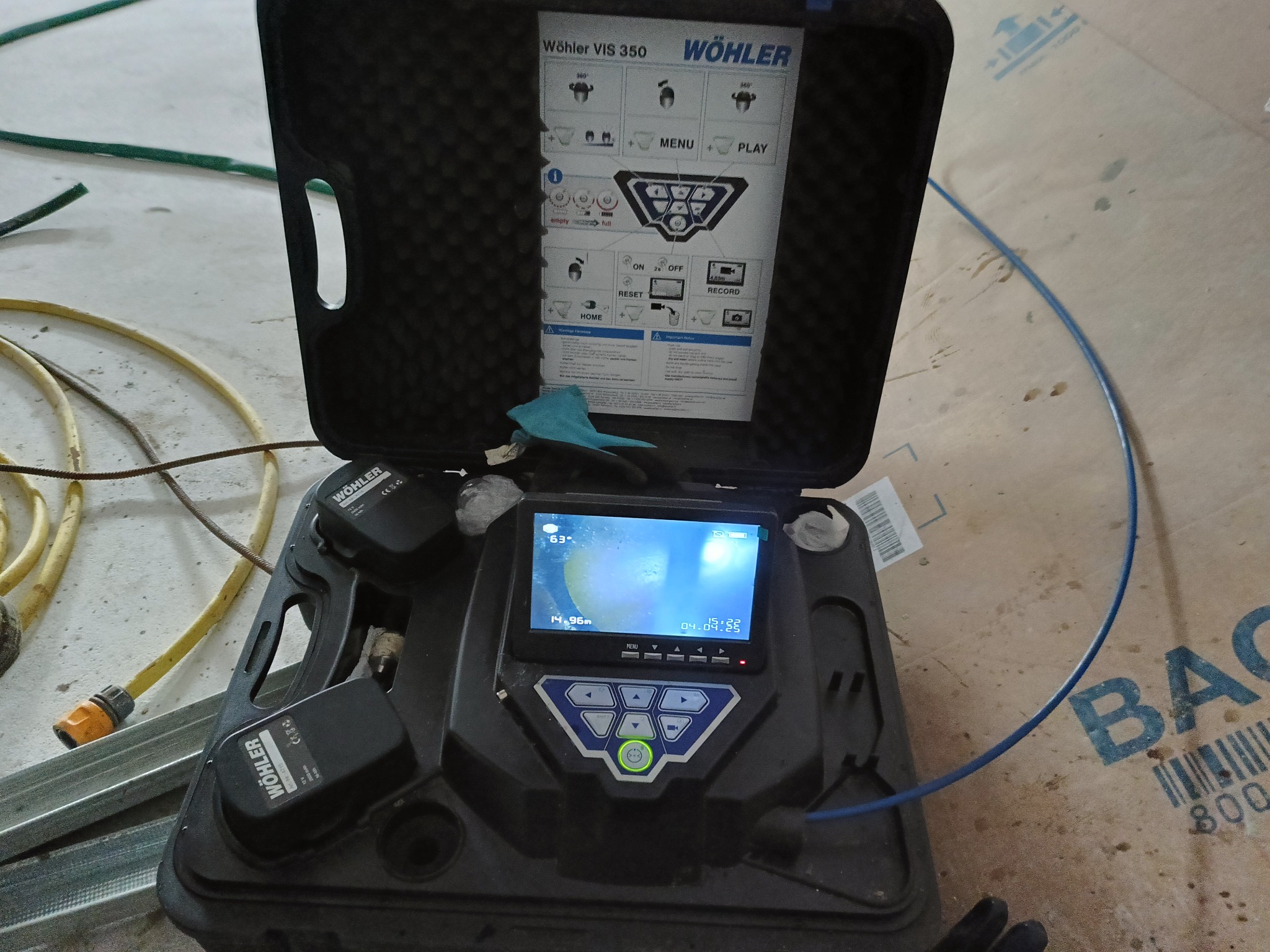 Kamera inspekcyjna Wöhler VIS 350 w otwartym futerale, pokazująca obraz z wnętrza rury na ekranie. Obok węże i profile metalowe.