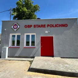 Nowo wybudowany budynek OSP w Starym Polichnie z czerwonymi drzwiami wejściowymi i białymi oknami, widok z zewnątrz w słoneczny dzień.