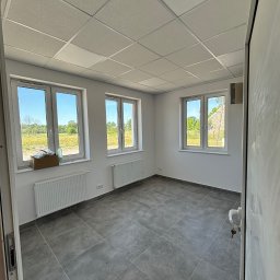 EFECT SP. Z O.O. - Wnętrze pustego pomieszczenia z trzema białymi oknami, szarą podłogą z płytek, białymi ścianami i grzejnikami, widok na zewnątrz przez okna na zielone pole i drzewa, oraz częściowo widoczne drzwi...