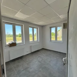 Wnętrze pustego pomieszczenia z trzema białymi oknami, szarą podłogą z płytek, białymi ścianami i grzejnikami, widok na zewnątrz przez okna na zielone pole i drzewa, oraz częściowo widoczne drzwi...