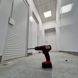 EFECT SP. Z O.O. - Wiertarka akumulatorowa na szarych płytkach podłogowych, na tle białych ścian i drzwi, w tym jednych z żaluzjami.