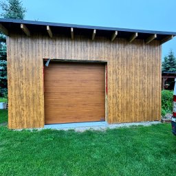 EFECT SP. Z O.O. - Nowo wybudowany garaż z drewnianą elewacją i segmentową bramą w kolorze drewna, częściowo widoczny samochód dostawczy na trawniku.