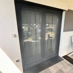 EFECT SP. Z O.O. - Zamknięta, plisowana moskitiera w czarnej ramie, zamontowana w drzwiach tarasowych, widok z zewnątrz, na szarej kostce brukowej.