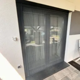 Zamknięta, plisowana moskitiera w czarnej ramie, zamontowana w drzwiach tarasowych, widok z zewnątrz, na szarej kostce brukowej.